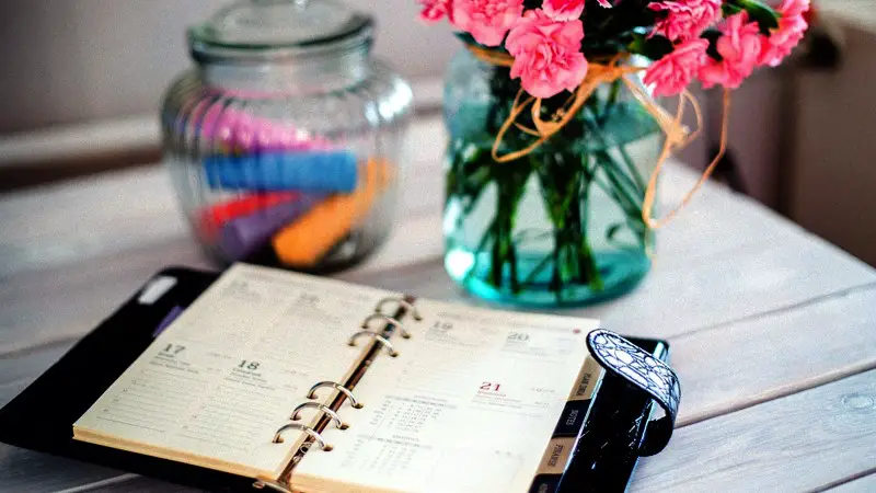 A paper planner open, and resting on a wooden desk next to some flowers and a container with colorful chalk.