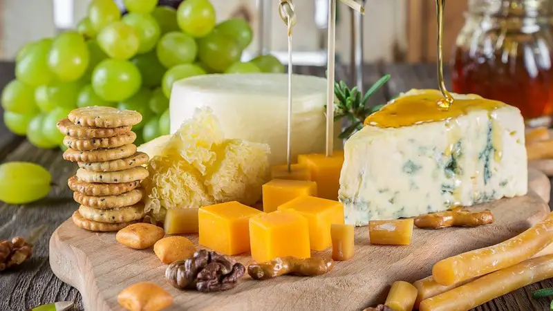 A cutting board full of cheeses, some nuts, and crackers, with grapes in the background.