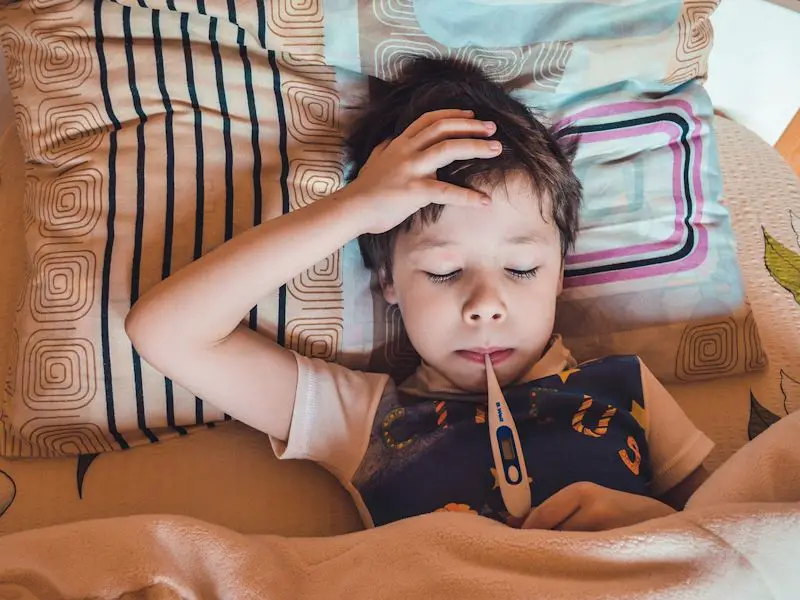 A boy laying down in bed getting his temperature taken for a fever. He has his hand on his forehead and does not seem to feel good.