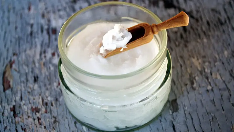 A jar of coconut oil in solid form, with a tiny wooden scoop, scooping up some of the oil.