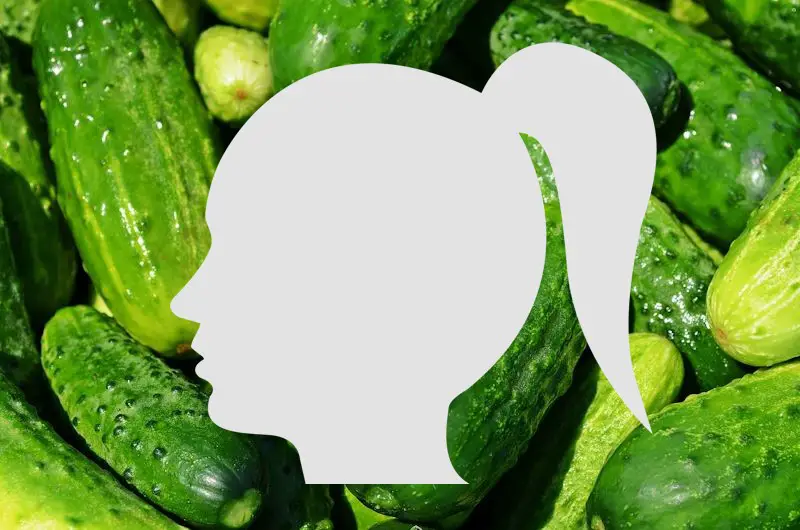 A silhouette of a woman's face and pony tail. This shadow is over a pile of cucumbers in the background.