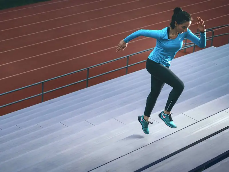 Woman in fitness gear running up the bleacher stairs at a track.