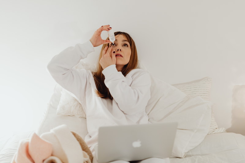 woman putting in eye drops while in bed and on her laptop