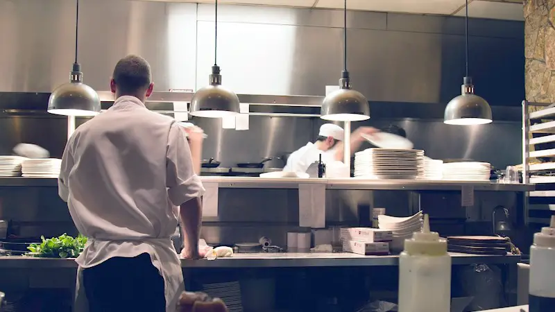 server in the kitchen of a busy restaurant waiting on the food food his table.