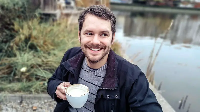 A man outdoors, smiling as he drinks his foamy coffee while sitting next to a body of water.
