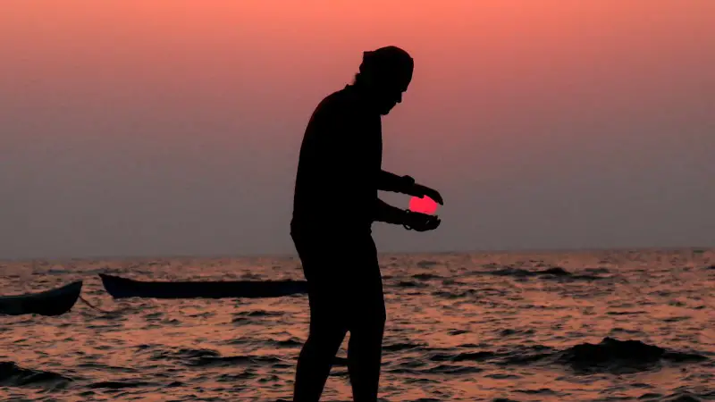 A man at the beach during sunset with his hands apart like he is holding the sun.