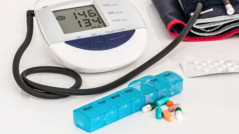 Medical blood pressure machine and cuff with a weekly pill container in front of it. The Friday pill flap is open with seven pills on the table in front of it.
