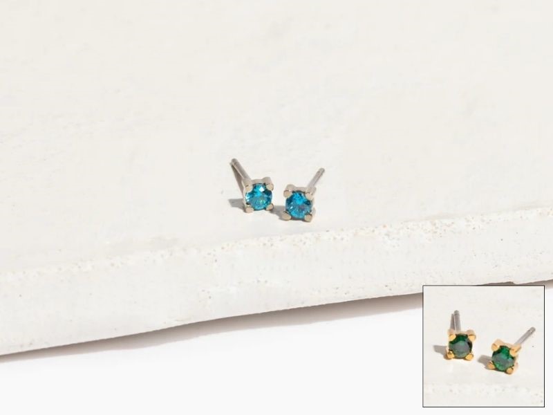 Two pairs of mini celeste titanium stud earrings with gemstones. The silver pair contains blue gems, and the gold pair contains green gems.