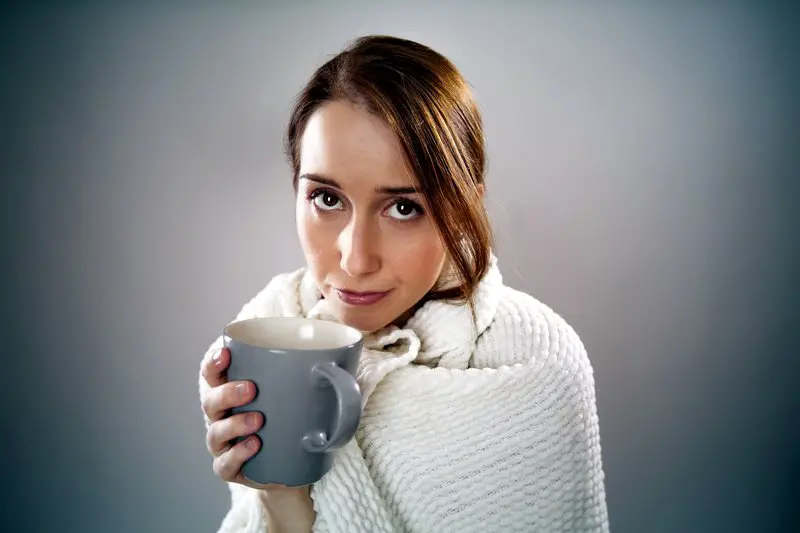 woman drinking tea while sick and wrapped in a blanket