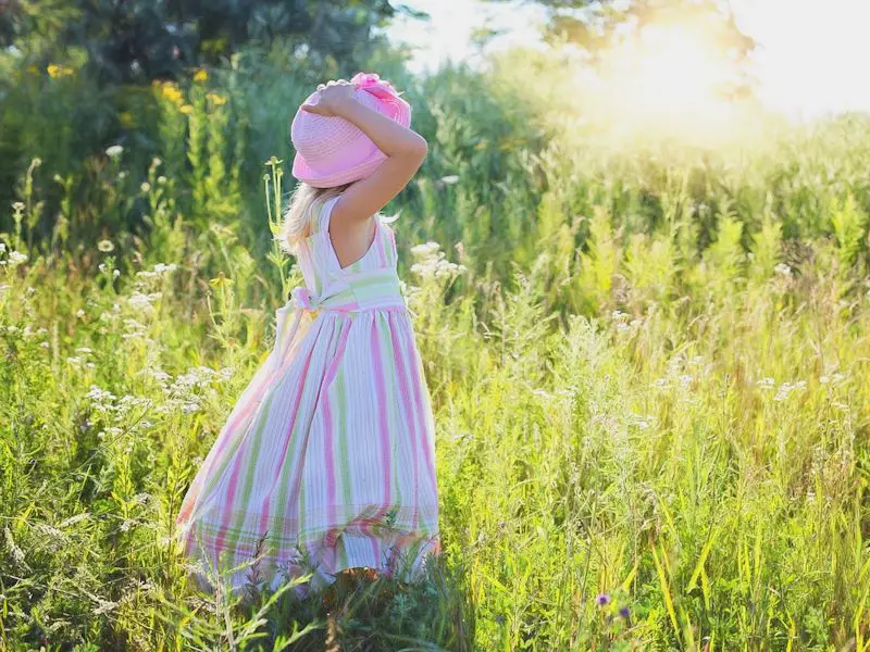 A little girl holding her hat on while walking through a field of tall grass in the summer.