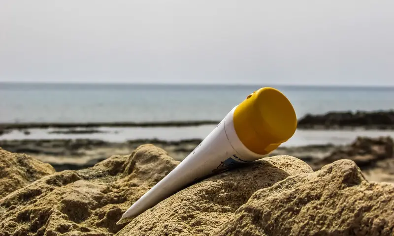 A squeeze tube bottle of sunscreen propped up in the sand at a beach, with water in the background.