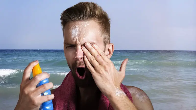 A man applying spray sunscreen at the beach, but he got it directly into his eyes.
