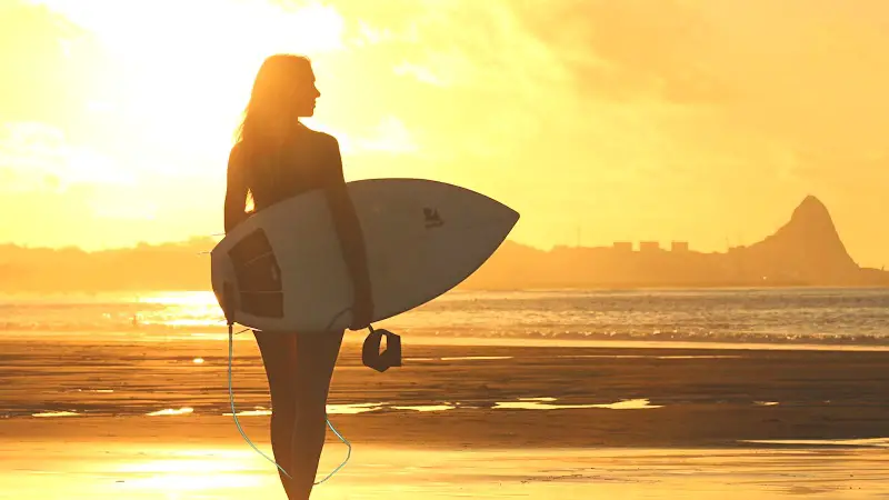A surfer woman walking on the beach with her surf board with an intense sunset in her background.