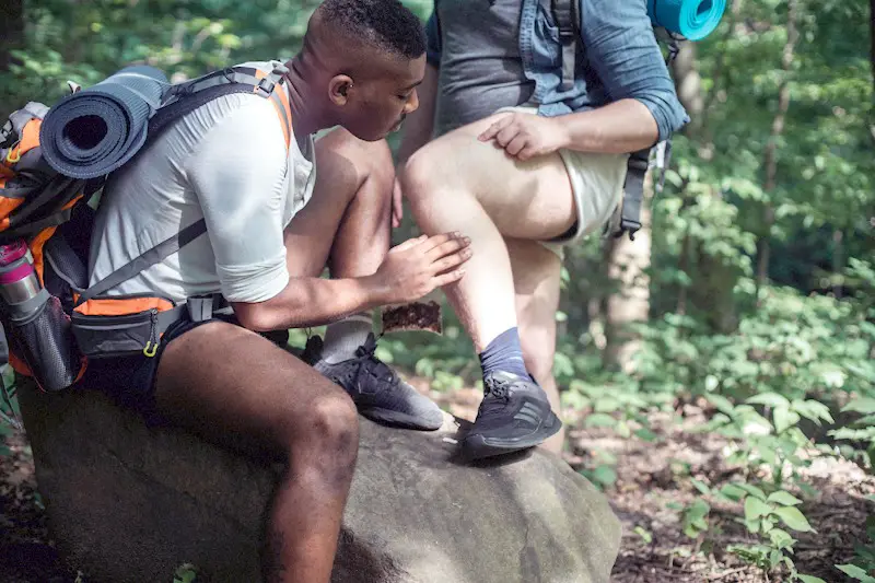 A person conducting a tick check on the leg of his hiking friend in the woods.