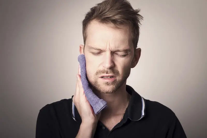 A man in pain holding a warm washcloth to his face. His pain is being caused by his tooth.