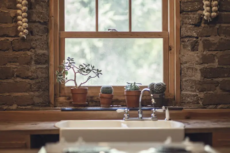 A shut wooden framed window in a kitchen with cactus on the sill.