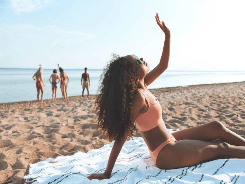 Woman with curly hair, laying on a towel at the beach with her hand in the air feeling the sun. There are four people in the background enjoying the sand and water.