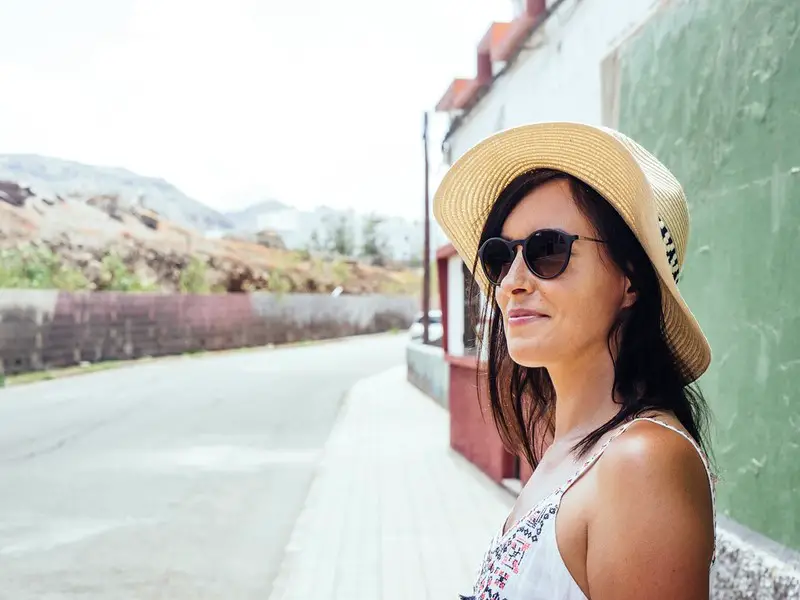 Woman looking across a street, wearing glasses, and a sun hat.