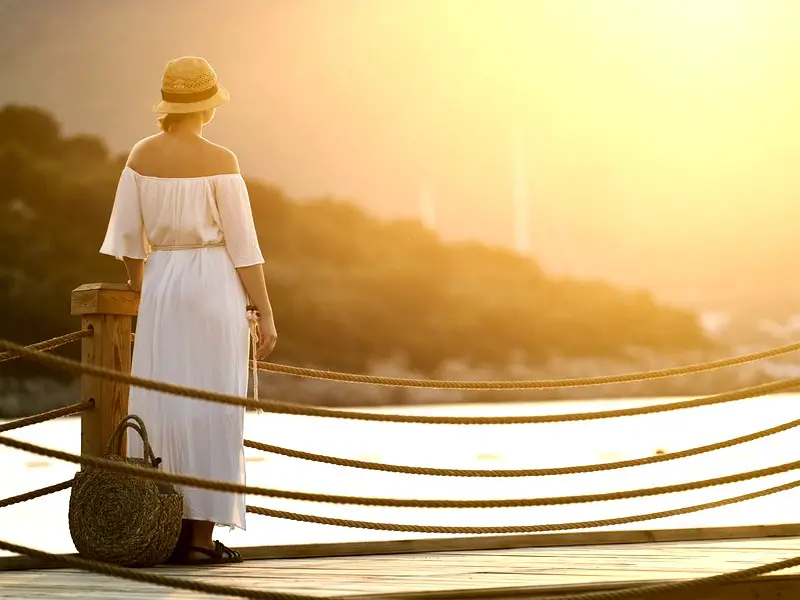 Woman waiting on a dock with rope rails during sunset. She is wearing a white dress and a pretty hat.