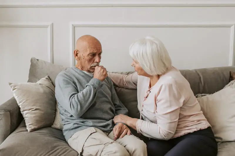 elderly man coughing on the couch with his spouse comforting him