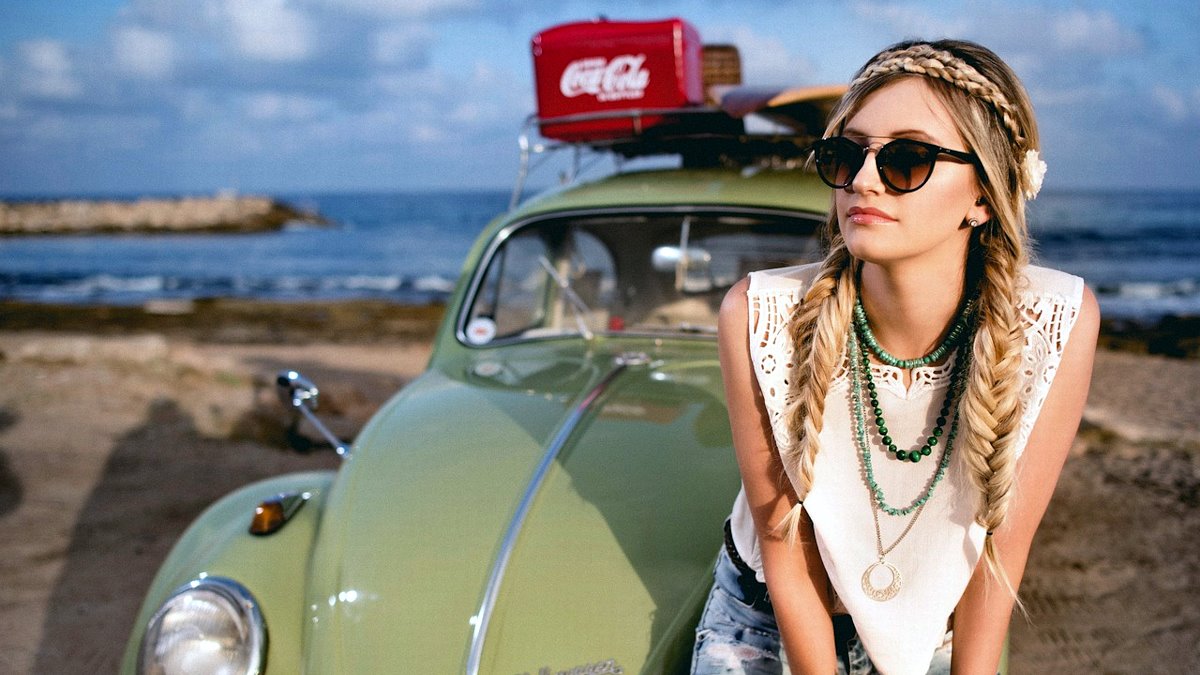 Woman sitting on the hood of her car, in sunglasses, while at the beach.