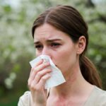Woman who is outdoors has really bad allergy symptoms. She is blowing her nose, has a neck rash, and watery eyes.